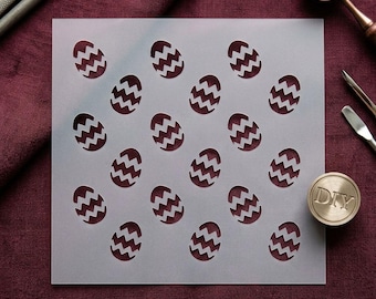 May include: A square, semi-transparent plastic stencil featuring a repeating pattern of egg shapes, each decorated with a horizontal zigzag line. The stencil is positioned on a dark red fabric surface alongside metal crafting tools and a round stamp.