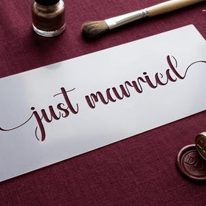 Könnte beinhalten: Eine rechteckige weiße Schablone mit dem Schriftzug "just married" in fließender Schreibschrift. Die Schablone ist umgeben von einem kleinen Glas mit Metallic-Farbe, einem Holzpinsel, zwei Spulen mit dünnem Silberdraht und einem Messingsiegel.