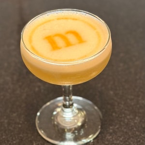 K&ouml;nnte beinhalten: Ein Cocktail in einem Coup&eacute;-Glas mit einem gelben Schaum oben. Der Schaum hat ein stilisiertes "M"-Design.