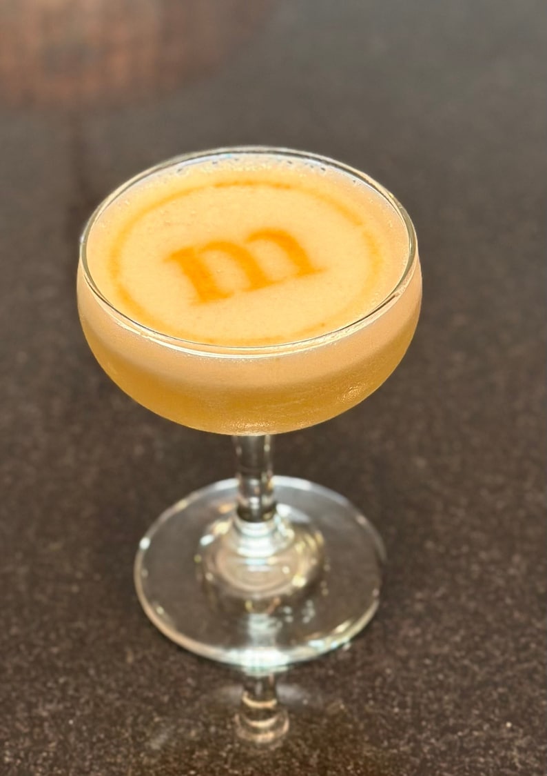 K&ouml;nnte beinhalten: Ein Cocktail in einem Coup&eacute;-Glas mit einem gelben Schaum oben. Der Schaum hat ein stilisiertes "M"-Design.