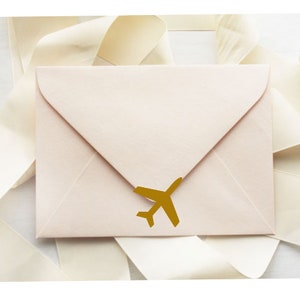 Könnte beinhalten: Ein blassrosa Umschlag mit einem goldenen Flugzeugaufkleber auf der Vorderseite. Der Umschlag ist von einem weißen Satinband umgeben.
