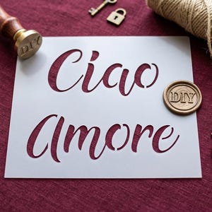 May include: A white stencil with the words "Ciao Amore" cut out in a cursive font. A DIY wax seal, keys, and a small padlock are scattered around the stencil on a burgundy surface. A wooden handled stamp is in the upper left corner.