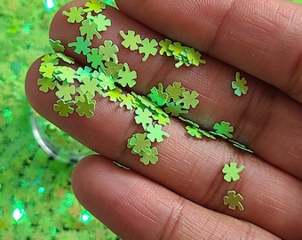 May include: A close-up of a hand holding green glitter shaped like four-leaf clovers. The glitter is scattered on the hand and on a surface below.