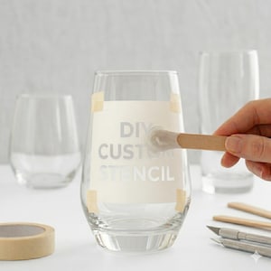 May include: Clear stemless wine glass with a stencil reading "DIY CUSTOM STENCIL" taped to the front. A small wooden brush is applying paint to the stencil. Two other glasses are in the background.