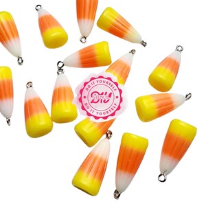 Könnte beinhalten: Eine Sammlung von 14 Candy-Corn-Anhängern mit silbernen Sprungringen. Die Anhänger sind aus weißem, orangefarbenem und gelbem Kunststoff und haben die Form von Candy Corn. Die Anhänger sind auf einem weißen Hintergrund verstreut.