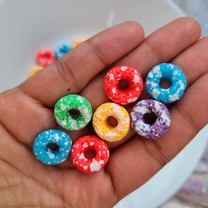 Könnte beinhalten: Eine Hand hält sieben bunte, donutförmige Müsli-Anhänger. Die Anhänger sind rot, orange, gelb, grün, blau, lila und weiß mit weißen Streuseln.