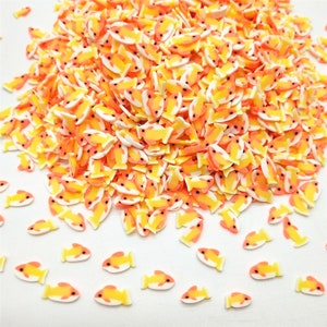 May include: A large pile of yellow and orange fish-shaped sprinkles on a white background. The sprinkles are small and have a white outline.