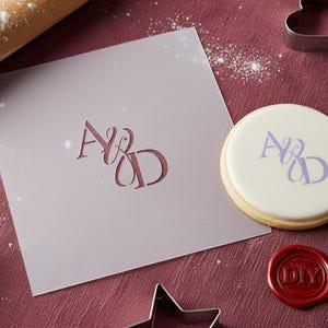 Op de afbeelding: Een lichtpaarse sjabloon met de letters "A&D" uitgesneden, een koekje met hetzelfde ontwerp en een rode zegel met de tekst "DIY". Een deegroller en koekjesvormen zijn ook aanwezig.