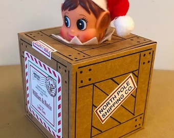 May include: A Christmas elf head wearing a red and white hat peeks out of a brown wooden crate. The crate has a "North Pole Shipping Co." sticker and a label that reads "William & Rosie".