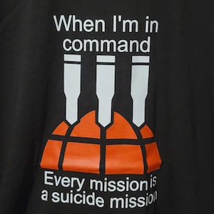 Puede incluir: Camiseta negra con el texto "When I'm in command" sobre un gráfico de tres misiles blancos sobre una forma naranja. Debajo del gráfico, el texto dice "Every mission is a suicide mission".