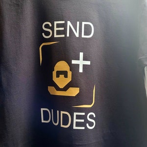 Puede incluir: Camiseta negra con la frase "SEND DUDES" en letras blancas. El diseño incluye un gráfico dorado de una cara y un signo más. La camiseta tiene cuello redondo y está hecha de un material suave.