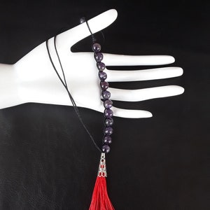 Pode incluir: Um colar de contas de ametista roxa com um tassel vermelho. O colar é encordoado em um cordão preto e tem um fecho prateado.