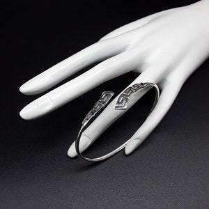 May include: A silver bracelet with a Greek key design. The bracelet is open and has a simple, elegant design.