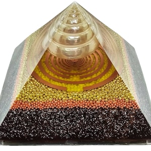 Op de afbeelding: Een heldere glazen piramide met een koperen spiraal en een heldere kristallen bol erin. De piramide is gevuld met lagen kleine gouden, koperen en zwarte kralen.