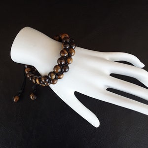 May include: A brown and gold beaded bracelet with a black cord tie. The bracelet is made of tiger's eye beads.