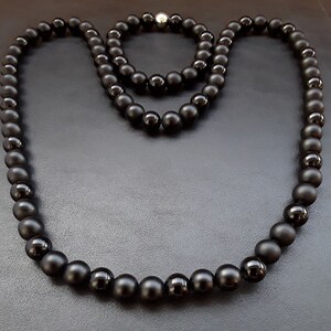 May include: Two black beaded necklaces, one longer than the other, on a black surface. The necklaces are made of matte black beads.
