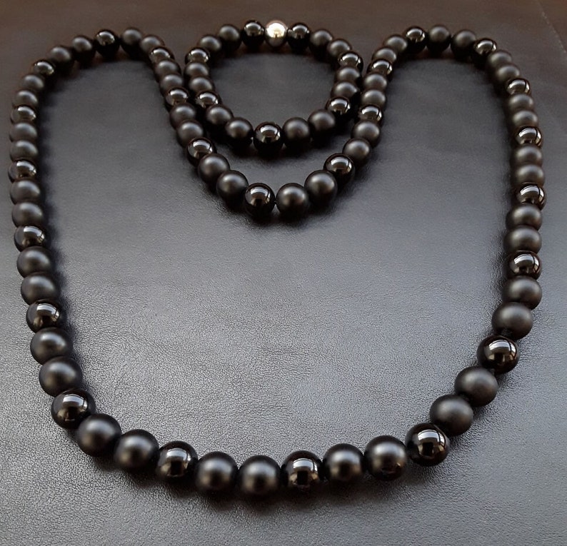 May include: Two black beaded necklaces, one longer than the other, on a black surface. The necklaces are made of matte black beads.