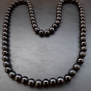 May include: A black beaded necklace with matte finish beads. The necklace is laid out in a U shape on a black surface.