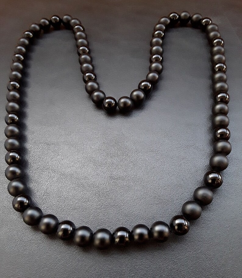 May include: A black beaded necklace with matte finish beads. The necklace is laid out in a U shape on a black surface.