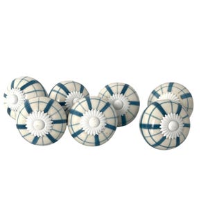 May include: A set of six round ceramic knobs featuring a cream-colored base with a dark blue grid pattern. Each knob is accented with a white, flower-shaped metal center piece that serves as a decorative detail on the front of the hardware.
