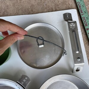 May include: A collection of vintage camping cookware, including a metal pot lid with a wire handle, a metal handle, and a metal plate. The lid is silver, and the handle is attached to the side. The items are on a white surface.