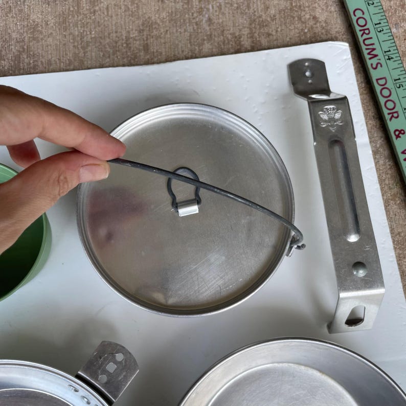 May include: A collection of vintage camping cookware, including a metal pot lid with a wire handle, a metal handle, and a metal plate. The lid is silver, and the handle is attached to the side. The items are on a white surface.