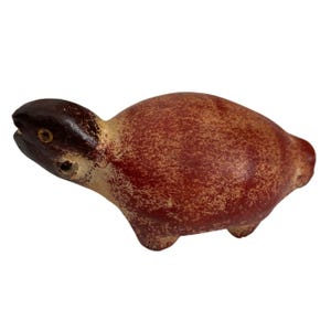 Puede incluir: Una pequeña figura de tortuga de cerámica pintada a mano con un esmalte rojo y marrón. La tortuga tiene la cabeza ligeramente levantada y un caparazón texturizado.