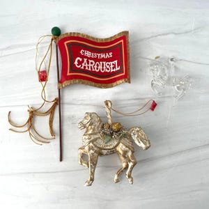 May include: A set of Christmas ornaments including a red flag with gold trim and the text "CHRISTMAS CAROUSEL" on a brown pole with gold tassels, a gold-painted horse ornament with a textured saddle, and a clear, transparent horse ornament.