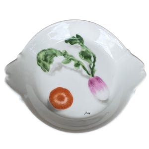 May include: White ceramic serving dish with a scalloped edge, featuring a hand-painted design of a radish and a carrot. The radish is purple and white, with green leaves. The carrot is orange. The dish is signed "J.V.M."