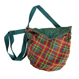 May include: A round, plaid shoulder bag with a green strap and flap. The bag features a red, green, and yellow plaid pattern. The flap is green with the text "Katie Wilkins" and a button closure. The strap is a solid green.