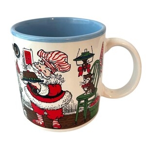 May include: A ceramic mug with a light blue interior and rim, featuring a festive illustration of Santa Claus wearing a red and white striped hat, holding a tray of cookies, alongside a small cat sitting on a green chair with a red bow.
