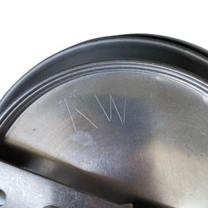May include: Close-up of a metal lid with the etched initials "KW". The lid has a circular shape with a black rim and a brushed metal surface. The initials are etched in a simple, clean font.