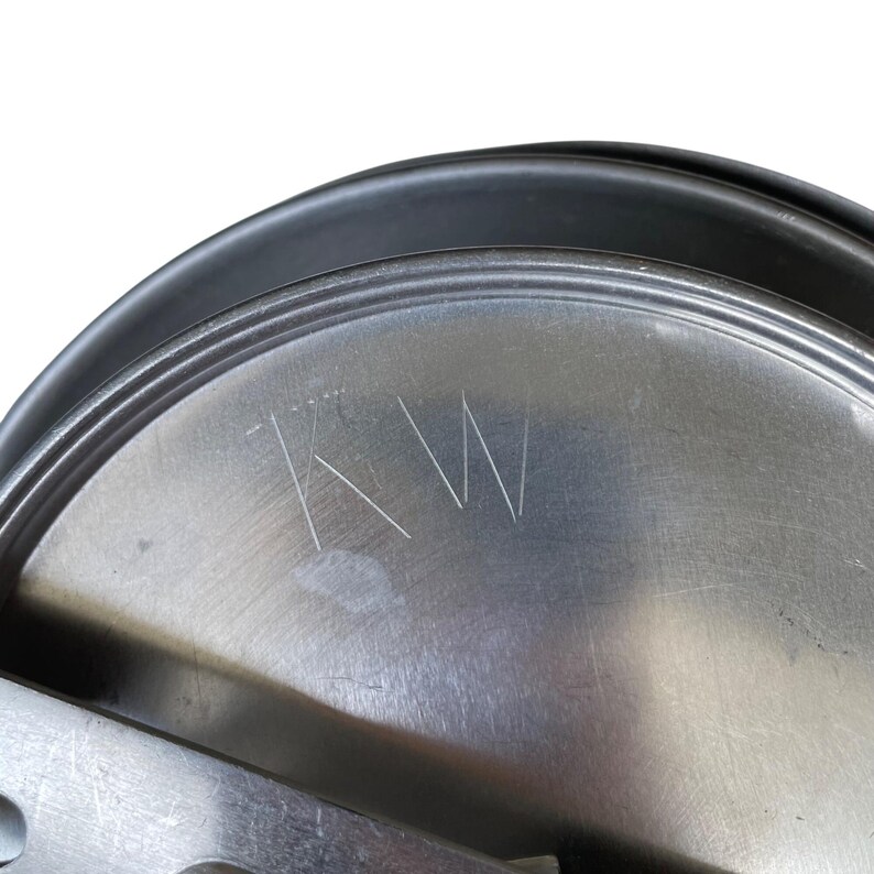 May include: Close-up of a metal lid with the etched initials "KW". The lid has a circular shape with a black rim and a brushed metal surface. The initials are etched in a simple, clean font.