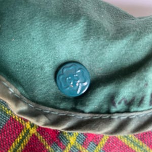 May include: Close-up of a green fabric hat with a teal button featuring a clover design. The hat has a red, yellow, and black plaid pattern on the brim. The hat is likely a vintage item.