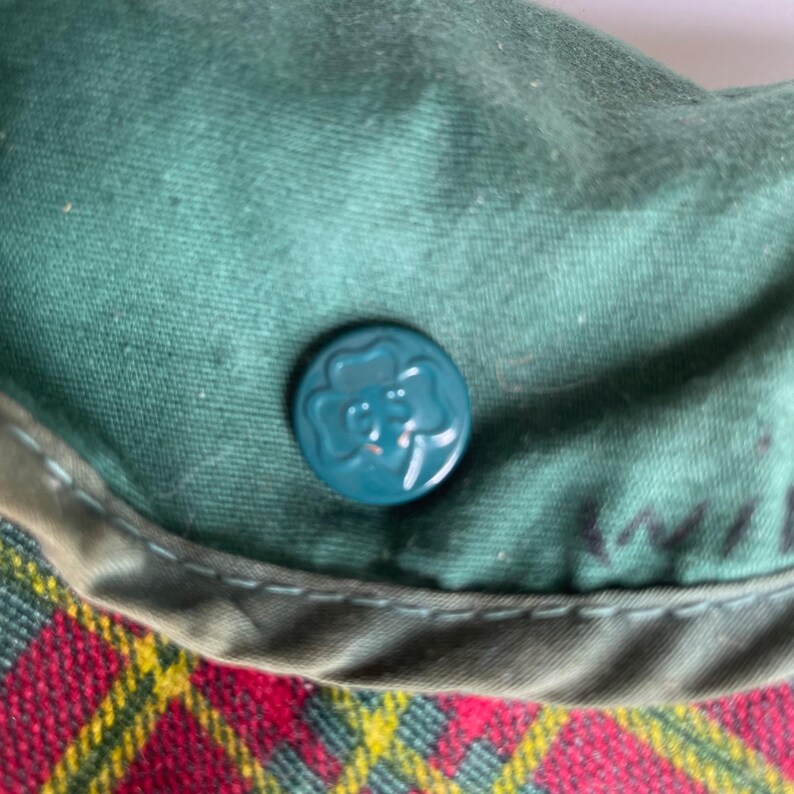 May include: Close-up of a green fabric hat with a teal button featuring a clover design. The hat has a red, yellow, and black plaid pattern on the brim. The hat is likely a vintage item.
