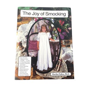 May include: A softcover instructional book titled The Joy of Smocking by Martha Pullen, Ph.D. The cover features a photograph of a child in a white smocked nightgown, surrounded by lace and fabric, with a list of contents including smocking designs.