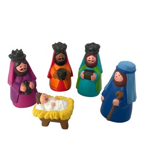 May include: A set of five small figurines including a baby in a yellow manger, a figure in a purple robe with a black crown, a figure in an orange robe with a black crown, a figure in a teal robe with a black crown, and a figure in a blue robe.