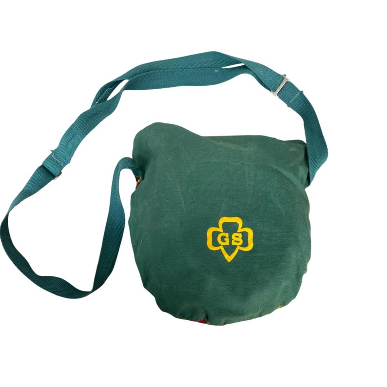 May include: A dark green shoulder bag with a long, adjustable strap. The bag has a round shape and features a yellow and gold Girl Scout emblem. The strap and bag are made of a durable fabric.