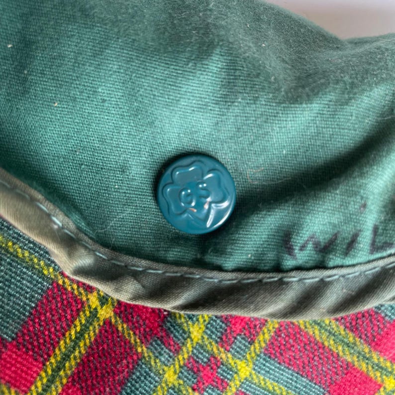 May include: Close-up of a green fabric with a teal button featuring a floral design. The fabric is part of a larger item, with a red and yellow plaid pattern visible at the bottom. The item appears to be vintage.