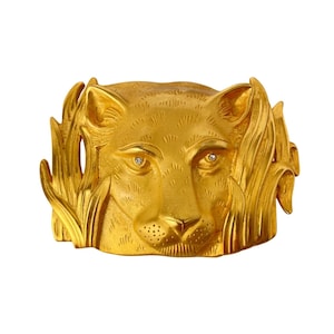 May include: A gold-coloured cuff bracelet with a detailed lion's head design. The lion's face is central, with two small, sparkling stones for eyes. The bracelet is surrounded by stylised foliage.
