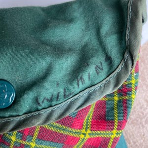May include: Close-up of a green fabric item with the name "Wilkins" handwritten in black. The item has a red, yellow, and green plaid pattern on the bottom. A teal button with a floral design is visible.