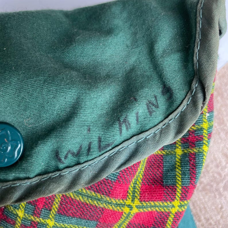May include: Close-up of a green fabric item with the name "Wilkins" handwritten in black. The item has a red, yellow, and green plaid pattern on the bottom. A teal button with a floral design is visible.