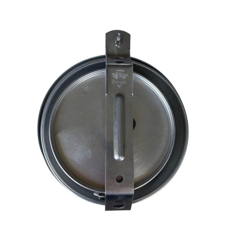 May include: A round, silver-colored metal object with a black rim and a silver bracket. The bracket has a decorative emblem and is attached to the top of the object. The object appears to be a lid or cover.