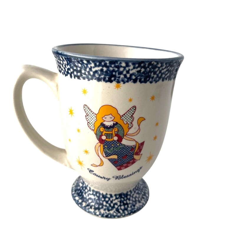 Pu&ograve; includere: Tazza in ceramica color crema con bordo e base maculati blu. La tazza presenta un'illustrazione colorata di un angelo che suona l'arpa, con le parole "Country Blessings" in basso. Stelle gialle sono sparse intorno all'angelo.