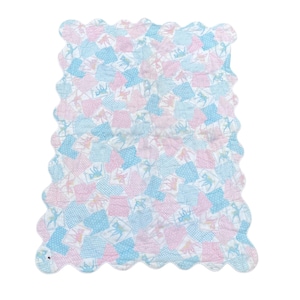 Puede incluir: Una manta acolchada con borde festoneado. La manta presenta un diseño de patchwork de cuadrados rosas y azules con un patrón repetido de bailarines. La manta es de un color pastel suave.
