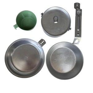May include: A camping mess kit with a green plastic cup, a metal pot with a lid, two metal pans, and a metal handle. The items are arranged on a white background.