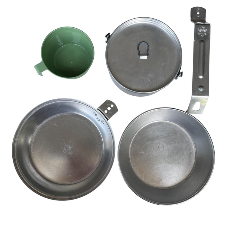 May include: A camping mess kit with a green plastic cup, a metal pot with a lid, two metal pans, and a metal handle. The items are arranged on a white background.