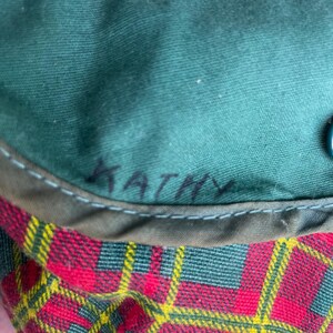 May include: Close-up of a green fabric with the name "KATHY" handwritten in black. The fabric is stitched to a red, yellow, and green plaid pattern.