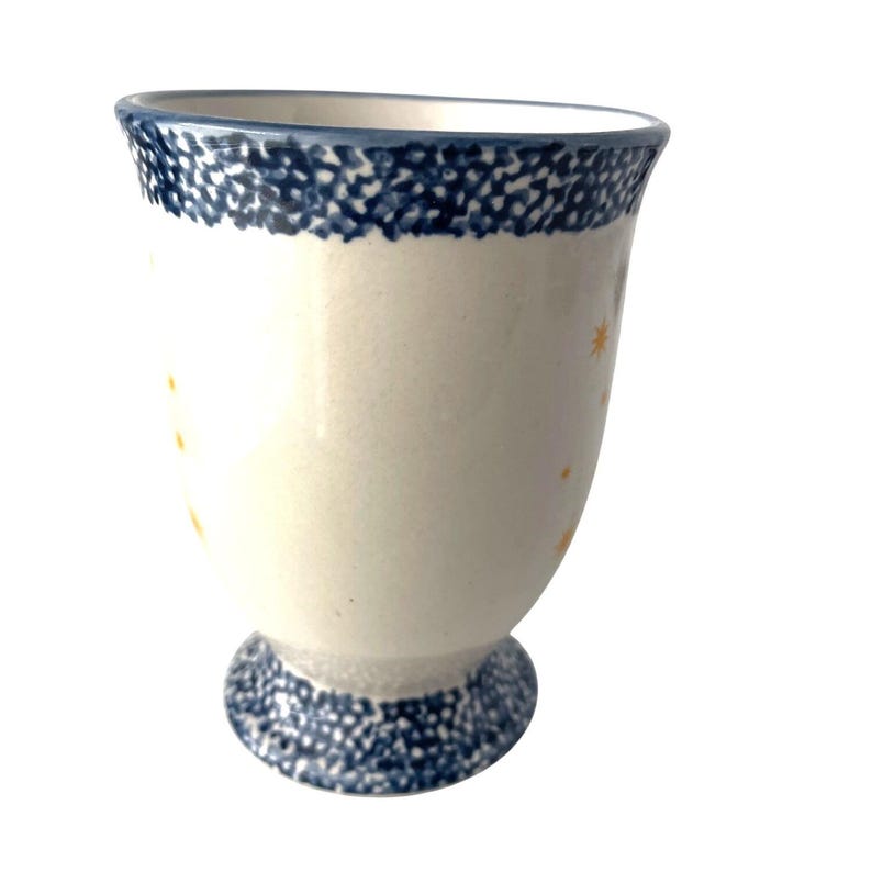 Pu&ograve; includere: Un calice in ceramica bianca con bordo e base maculati blu. Il calice ha un corpo color crema con piccoli disegni a stella gialla. Il calice &egrave; alto circa 13 cm.