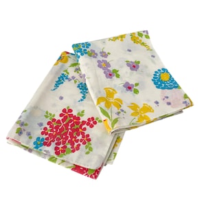 May include: Two white floral patterned handkerchiefs. One handkerchief features a cluster of red flowers, while the other displays yellow daffodils, blue and purple flowers. The handkerchiefs are folded and layered.